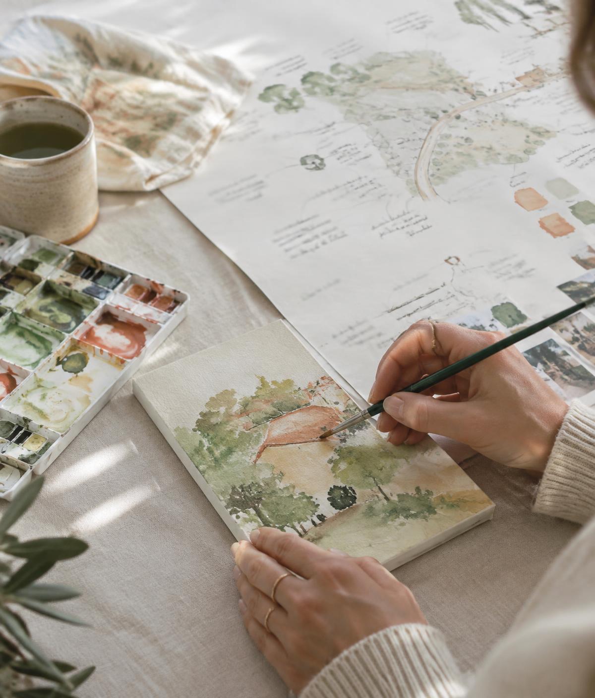 Mãos pintando aquarela sobre uma pequena tela, ao lado de um mapa de planejamento